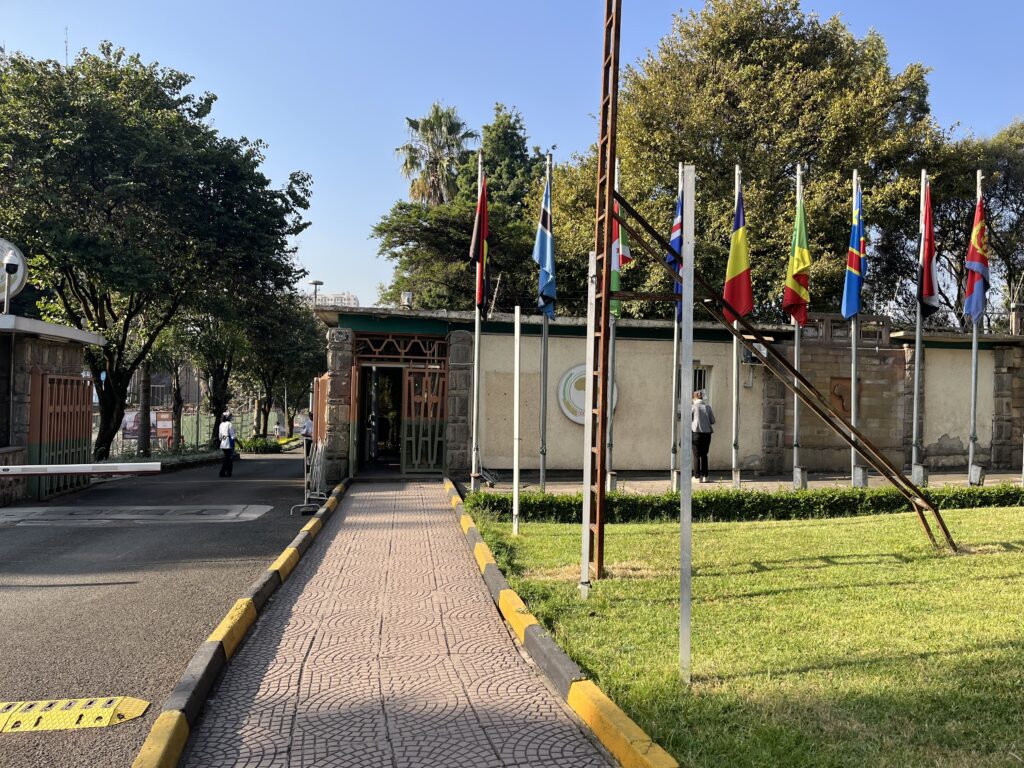 Picture of an entrance to the African Union compound. The picture shows a road on the left and a little footpath in the centre of the picture both leading up to a wall with a doorpost. On the right multiple flags of african states are visible.