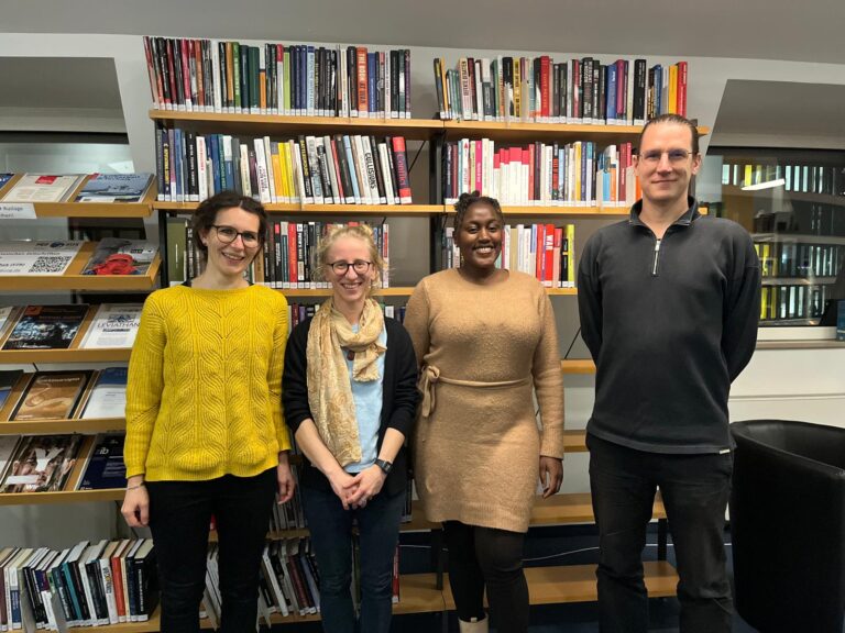 Four people standing in front of a book shelve at PRIF Library smiling to the camera.