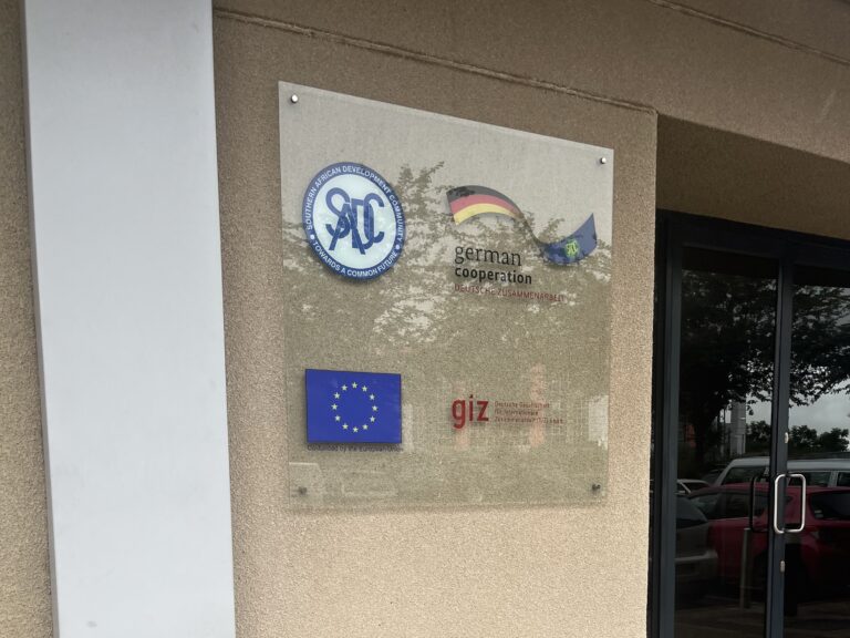 Picture shows a sign with the logos of SADC, German Cooperation, the EU, and the GIZ. It is a glass panel mounted to a wall.