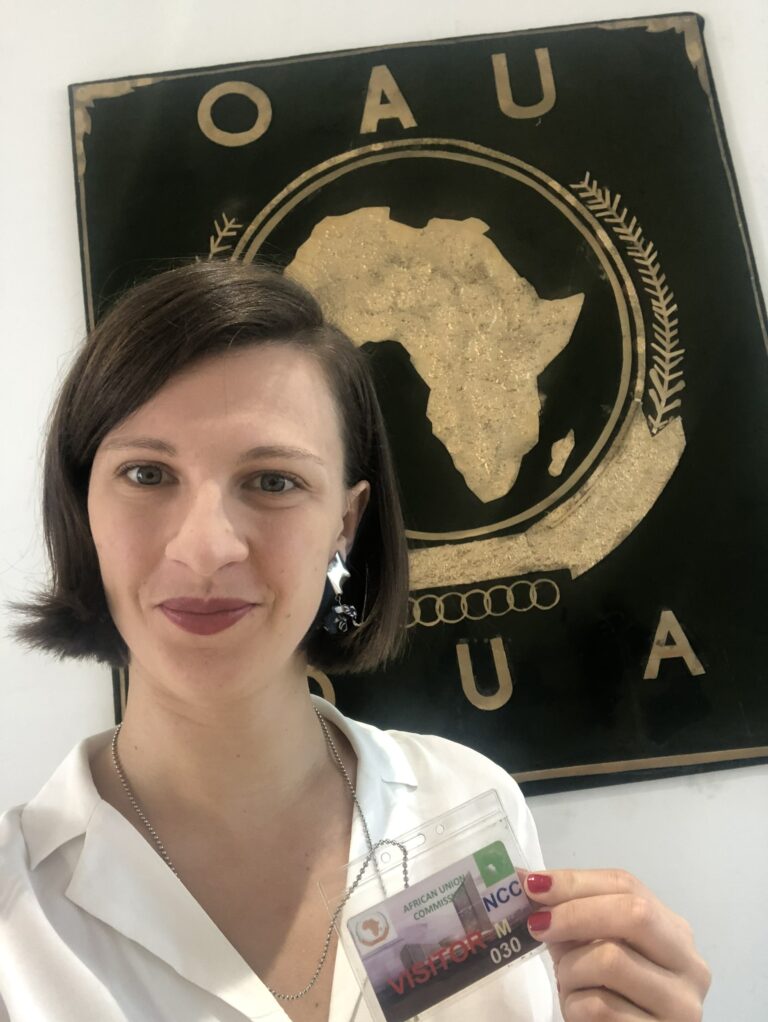 selfie of researcher Camille Vern. She is holding her visitor ID in front of a green-golden logo of the OAU and AU