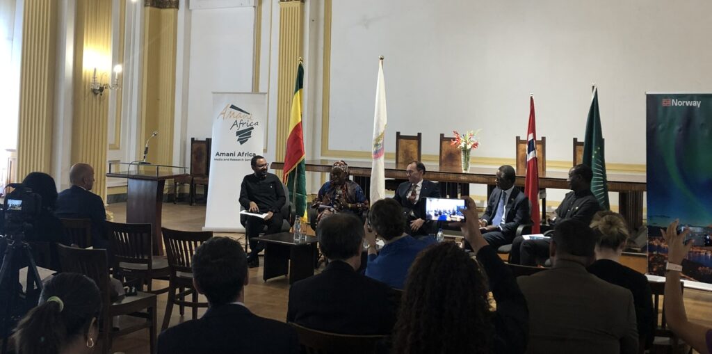 You can see a conference panel with five persons seated in front of a seated audience. The picture is taken from the audience.