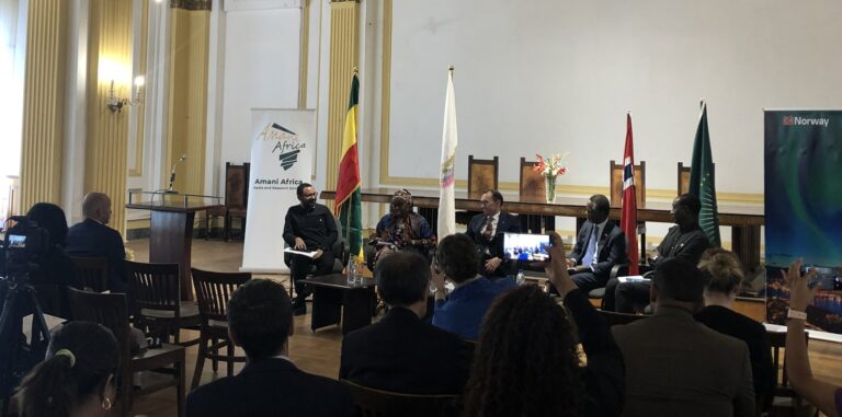 You can see a conference panel with five persons seated in front of a seated audience. The picture is taken from the audience.