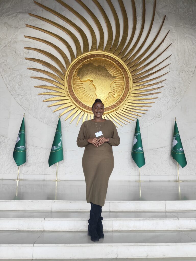 Thee Picture shows ANCIP Fellow Hilda Milka Koyier in front of the AU Logo and Flags.