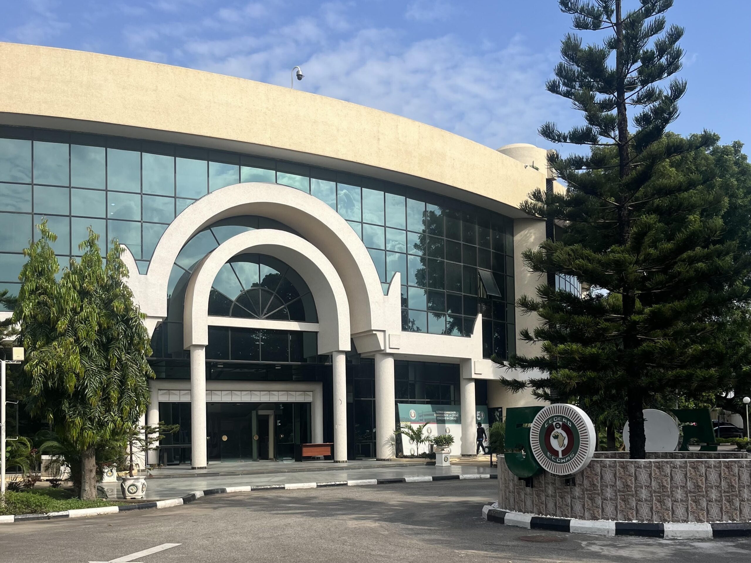 Picture shows the main entrance of the ECOWAS Headquarters on a sunny day. The building has a big glass front with an arched entry. In the front to the left is the ECOWAS Logo on a wall.