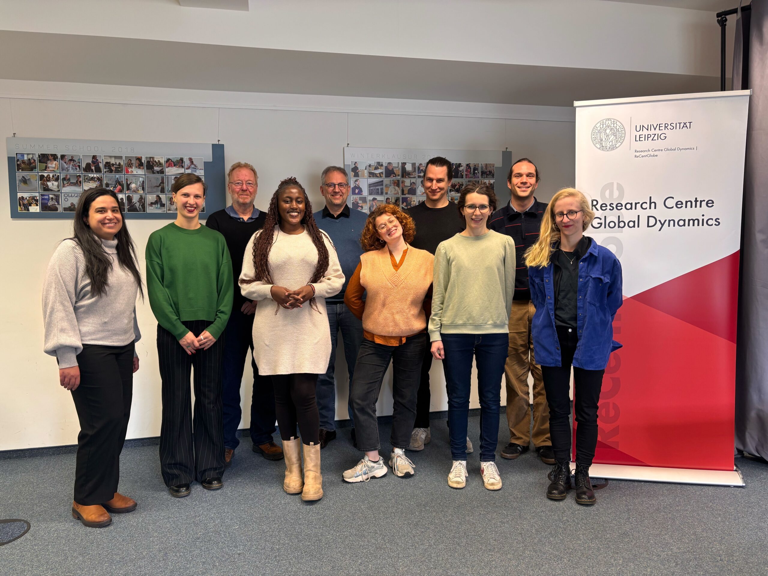The picture shows the ANCIP Project Members at a venue in the ReCentGlobe Institute, Leipzig University. The members are standing as group, smiling into the camera, with a banner of the ReCentGlobe Institute to the right.